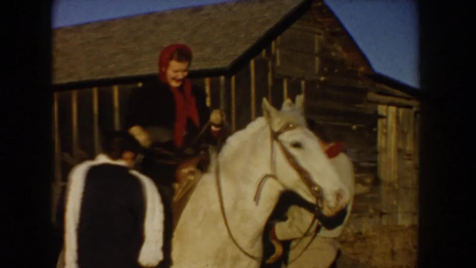 In 1955 Idaho ranch footage, a woman in a red headscarf sits on a white horse near a weathered wooden barn while another person stands close by.