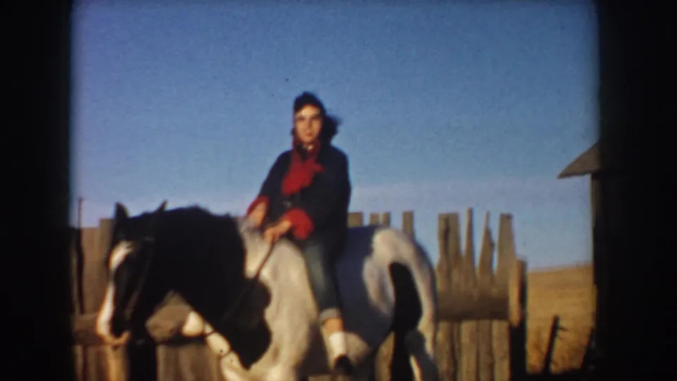 A woman in dark outerwear and a red shirt rides a black-and-white horse near a rough wooden fence in rural Idaho home-movie footage.