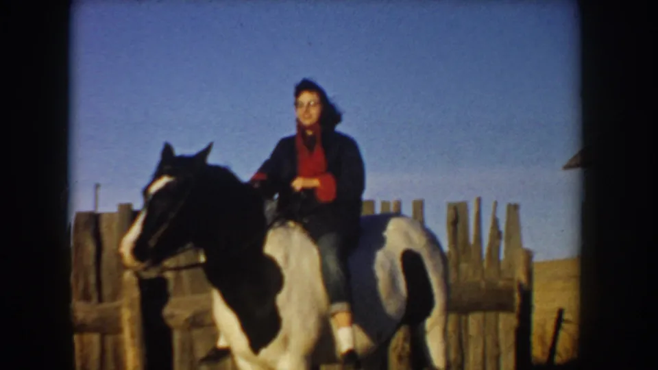 A woman on horseback pauses near a wooden fence in rural Idaho, framed by open grassland and clear sky in faded 1955 8mm color.