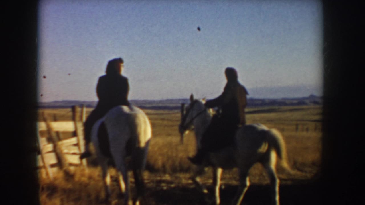 In 1955 Idaho home-movie footage, two riders on light horses move away from a wooden fence across golden prairie.