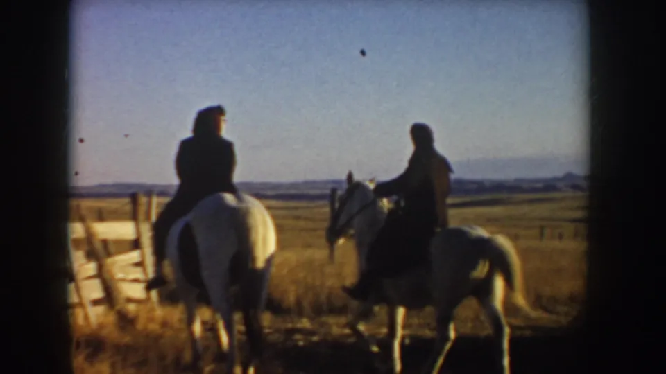In 1955 Idaho home-movie footage, two riders on light horses move away from a wooden fence across golden prairie.