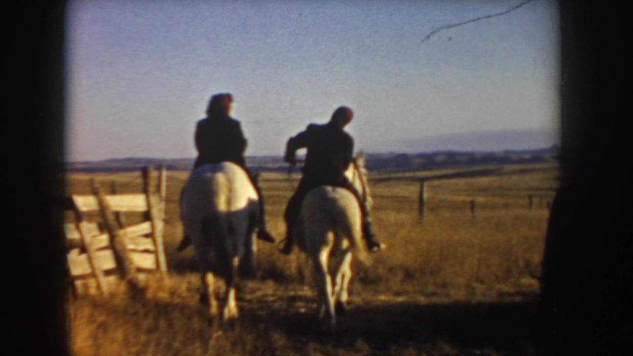 Two riders on horses moving away across dry grass beside a fence.