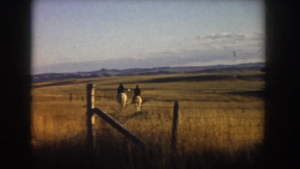 The riders appear smaller in the frame as they continue across the 1955 Idaho prairie beyond a foreground fence post.
