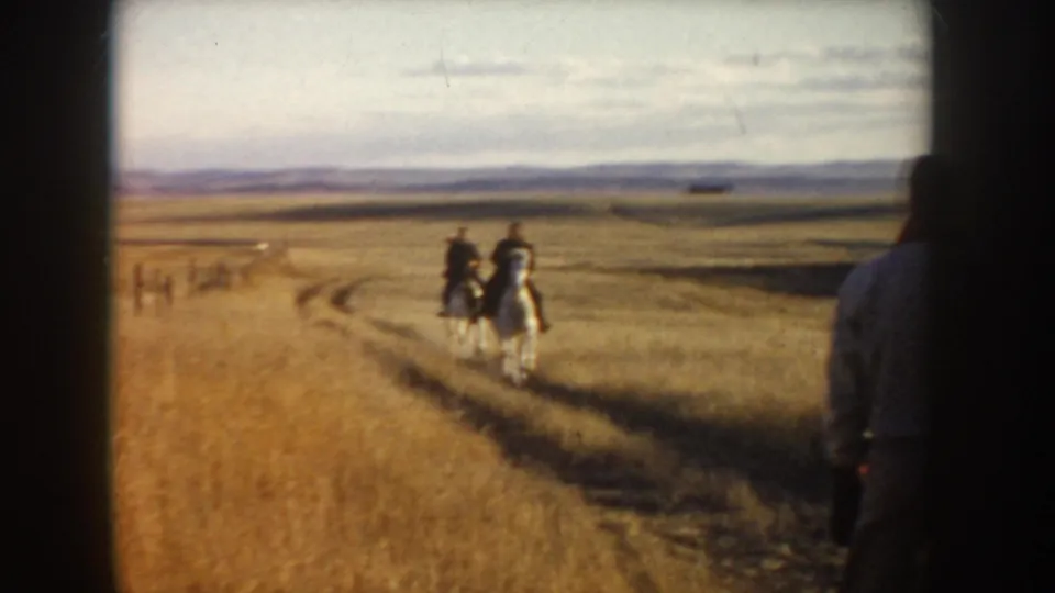 Two riders on light horses follow a pale dirt track through open Idaho grassland, with a partial figure at the right edge.