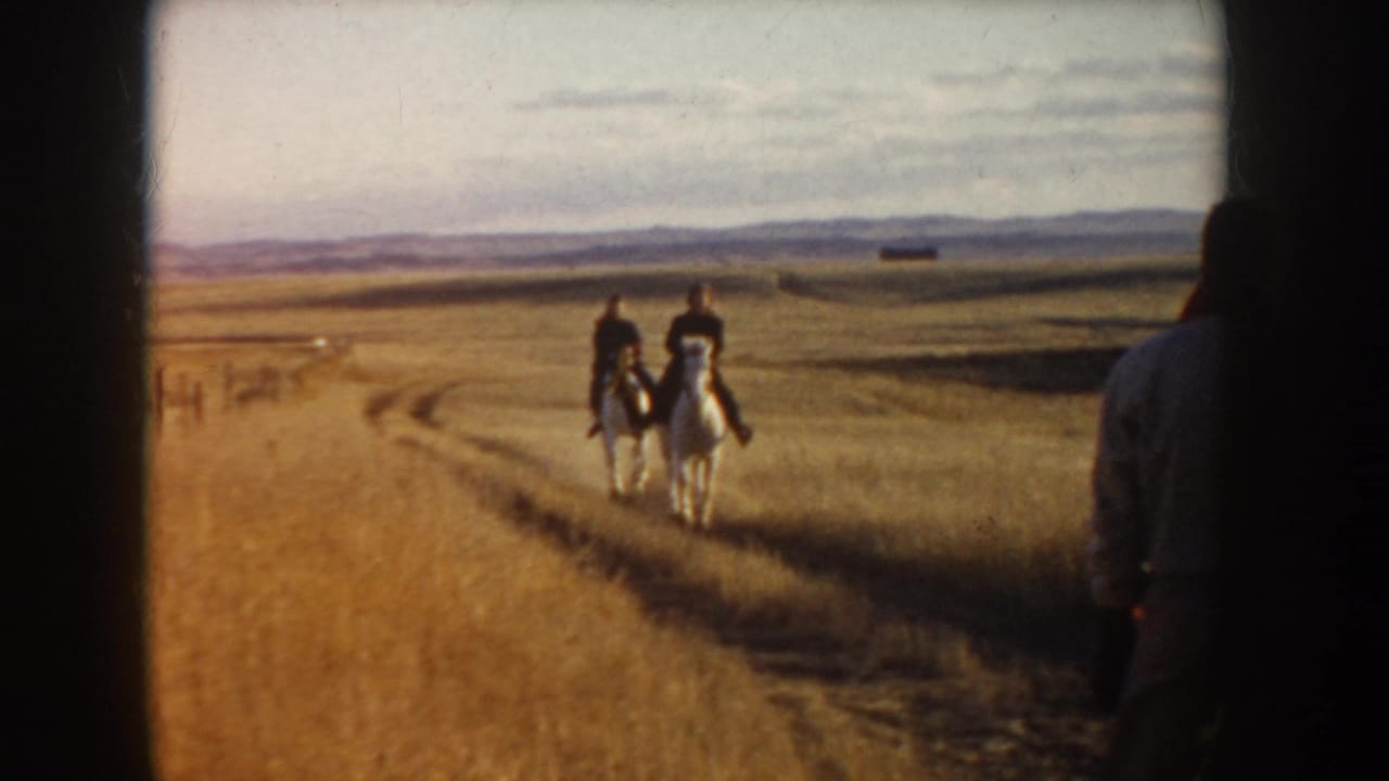 In 1955 Idaho 8mm footage, two riders on white horses move away across a dirt track in a wide rural landscape.