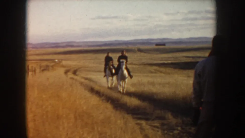 In 1955 Idaho 8mm footage, two riders on white horses move away across a dirt track in a wide rural landscape.