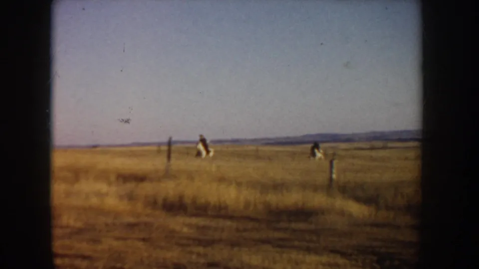 The riders appear small in the 1955 Idaho landscape, crossing dry grassland near scattered fence posts and a low horizon.