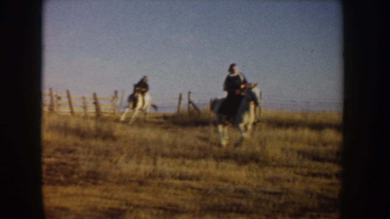 Two riders on horseback pass through dry grass near a fence.