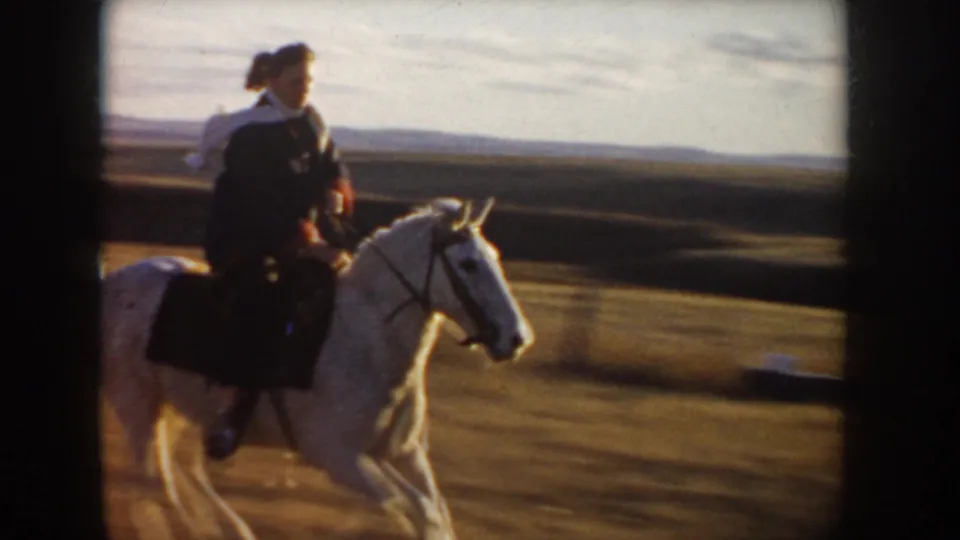 A woman in dark clothing and a light scarf rides a white horse through open Idaho countryside in 1955 footage.