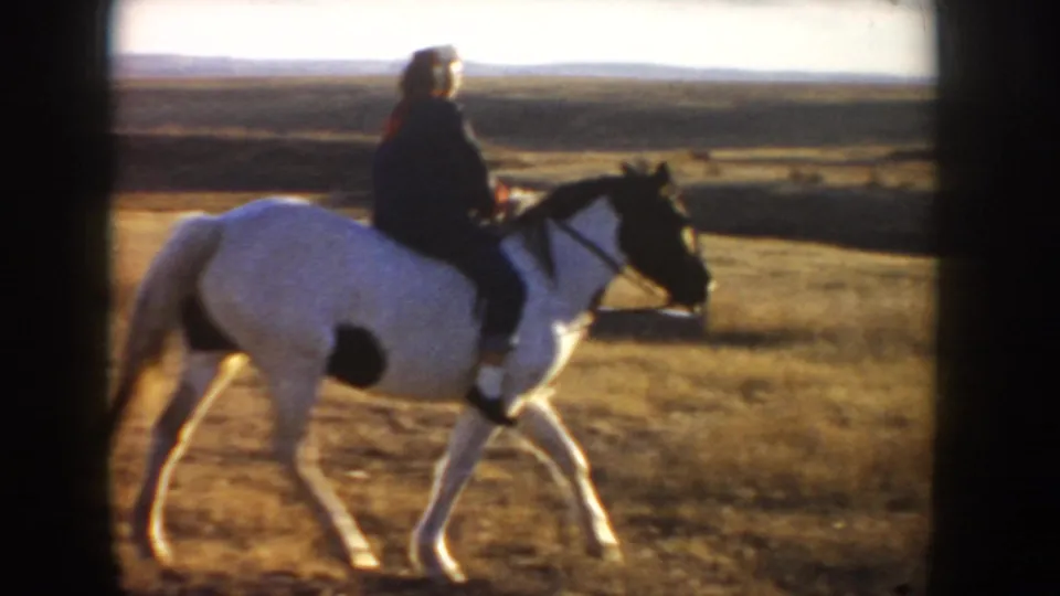 Side view from 1955 Idaho footage showing a rider on a black-and-white horse crossing sunlit rural grassland.