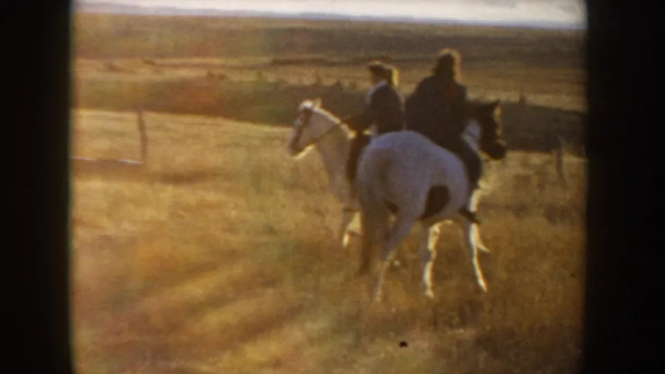 Wide 1955 Idaho home-movie frame with two riders on horses moving away across golden grassland in sunset light.