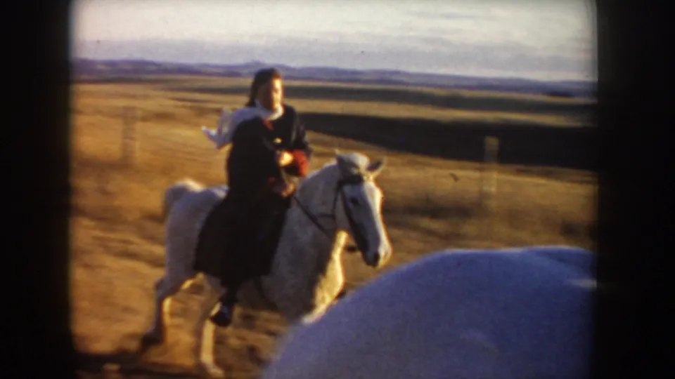 A woman in dark clothing and a light shawl rides a white horse through a rural Idaho field in 1955 footage.