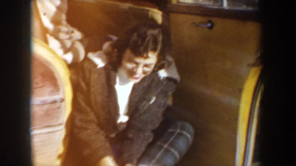 In a 1955 Idaho home-movie frame, a young woman smiles while seated inside a yellow mid-century car and adjusting footwear.