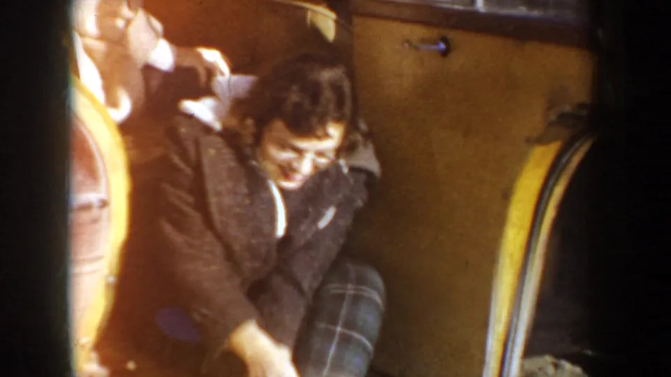 Young woman in a dark jacket and plaid pants smiles inside a 1950s Idaho car doorway, with the yellow door frame beside her.