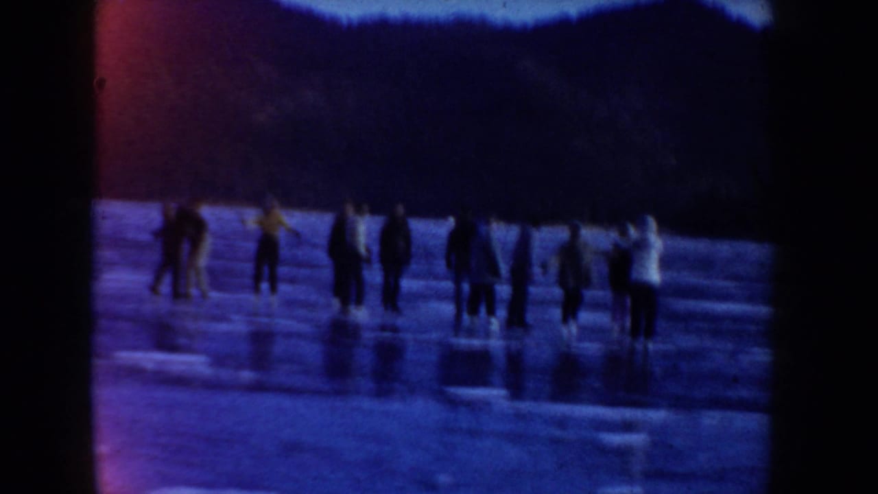 1955 Idaho frame showing several bundled skaters standing on lake ice below wooded hills at dusk or low light.