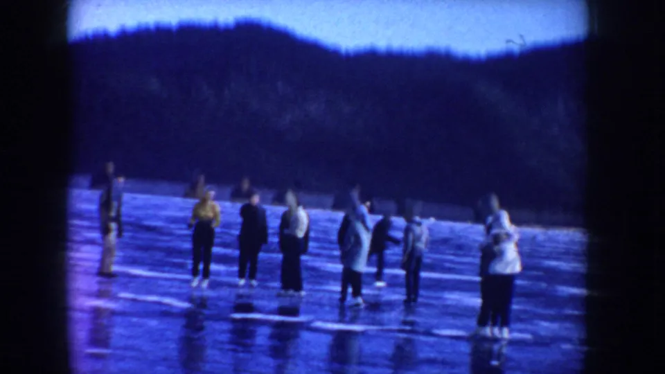 1955 Idaho home-movie frame of a group of skaters standing on reflective frozen lake ice with hills behind them.