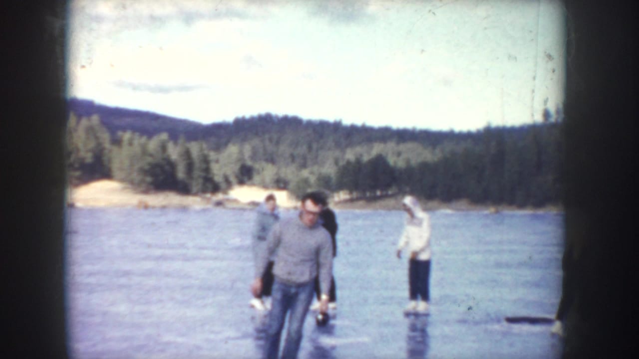 1955 Idaho home-movie frame showing several people skating cautiously on a frozen lake bordered by evergreen hills.