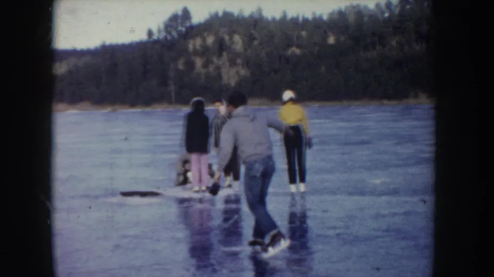 A man skates toward others on a frozen lake in Idaho, with wooded hills and soft 8mm color fading visible.