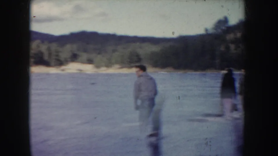 Home-movie frame of a man on a frozen Idaho lake, with other skaters near the edge and wooded hills beyond.