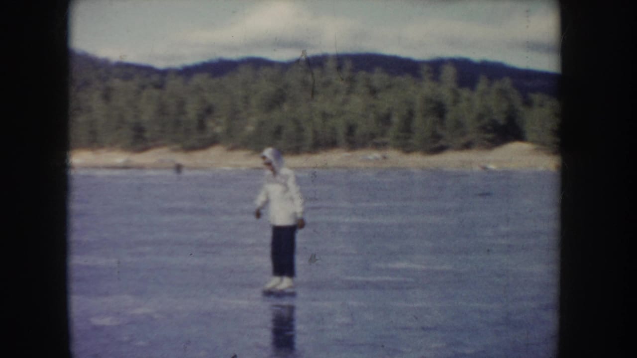 1955 Idaho 8mm frame showing an adult man skating alone on a frozen lake before a tree-lined shoreline and low hills.
