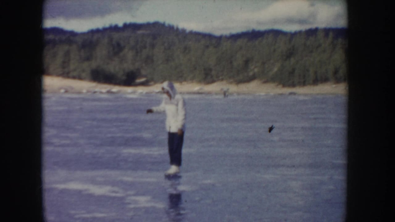 Man stands or glides on lake ice with one arm raised toward the wooded shoreline.