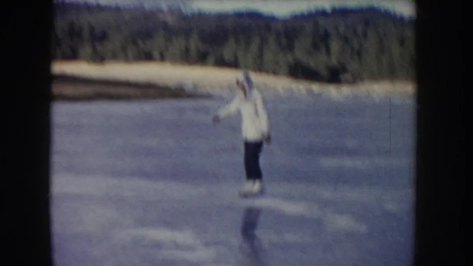 Soft 8mm frame of a man skating on a frozen Idaho lake, with motion blur, forested shoreline, and low hills visible.