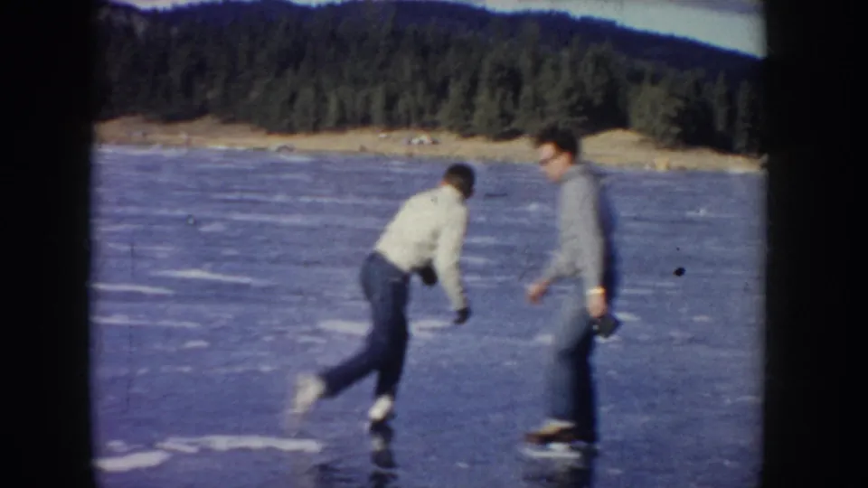Two men skate on a frozen Idaho lake, with one leaning into motion and another standing nearby before a wooded hillside.