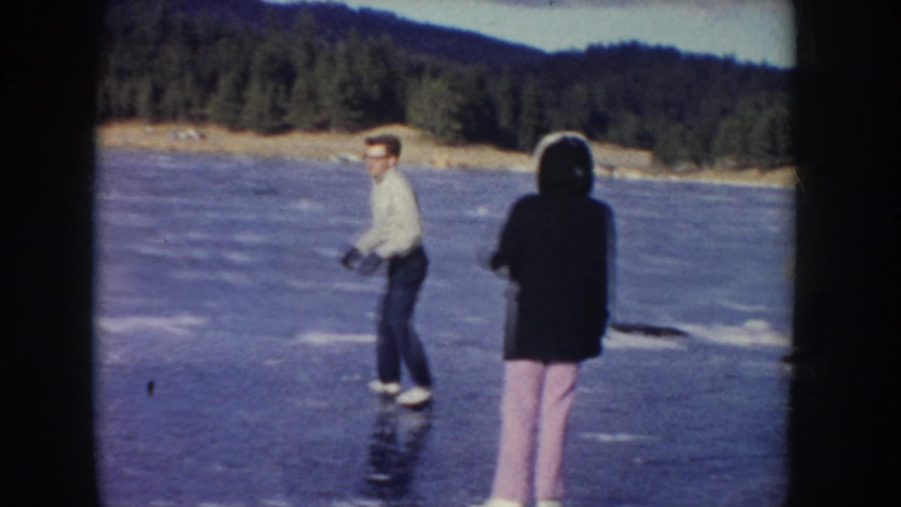 1955 Idaho home-movie frame showing adults ice skating on a frozen lake near a forested shoreline.