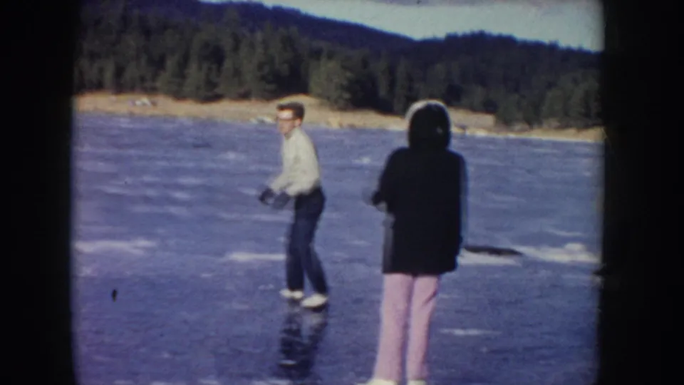 1955 Idaho home-movie frame showing adults ice skating on a frozen lake near a forested shoreline.