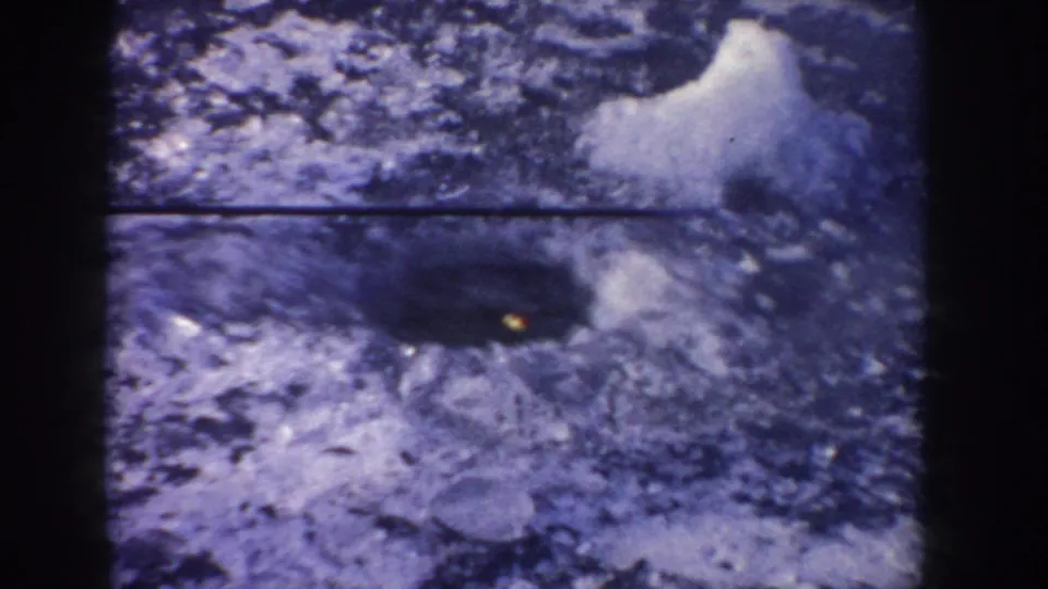 1955 Idaho home-movie frame of a dark fishing hole in snow-covered ice, with a small orange point near the opening.