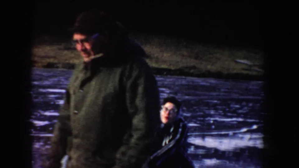 1955 Idaho 8mm frame showing two bundled people beside a frozen lake, with a pickup truck in the background.