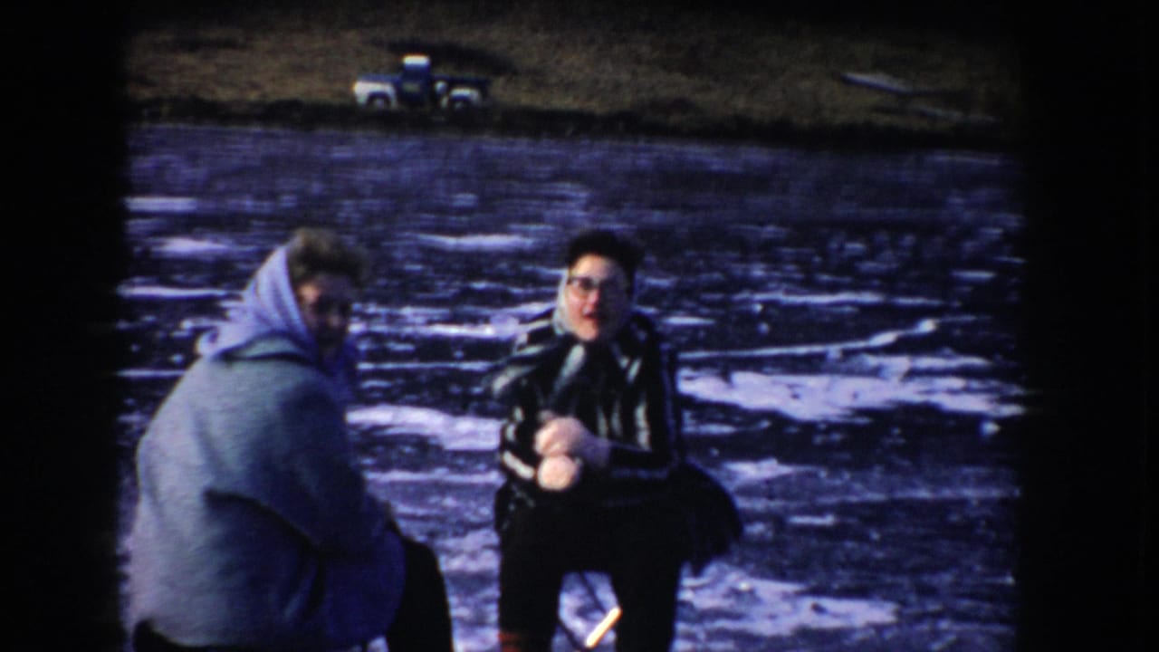 In 1955 Idaho home-movie footage, two adults in winter clothing sit on folding chairs on a frozen lake, with a vehicle visible on the far shore.