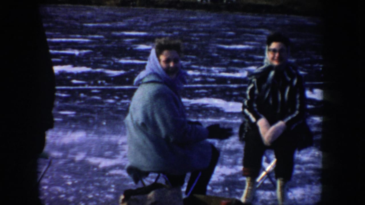 Two adults sit on folding chairs on the frozen lake in Idaho, one wearing a hooded coat and the other in dark winter clothing with skates visible.
