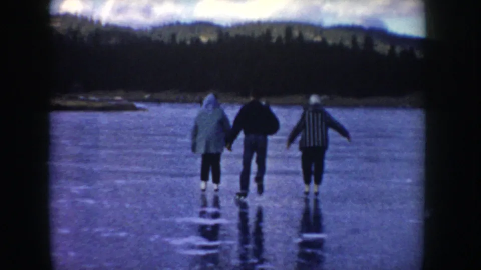 Three adults in winter clothing move away across the frozen Idaho lake, with their reflections visible on the ice and wooded hills in the distance.