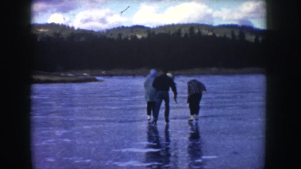 Three adults are seen from behind on the frozen Idaho lake, moving cautiously across the ice beneath a dark wooded shoreline and cloudy sky.