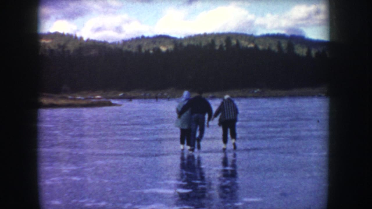 Three adults in winter clothing walk cautiously on reflective lake ice in Idaho, seen in soft 1950s 8mm footage.