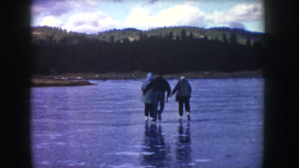 Three adults in winter clothing walk cautiously on reflective lake ice in Idaho, seen in soft 1950s 8mm footage.