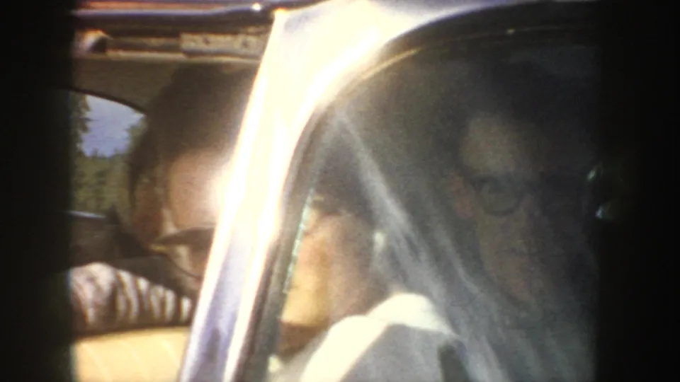 A close 1950s home-movie shot shows a person inside a car, partially obscured by window glare and soft focus.