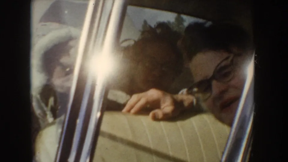 Two adults appear beside a car window in a candid 8mm travel shot, with glare and tight framing obscuring details.