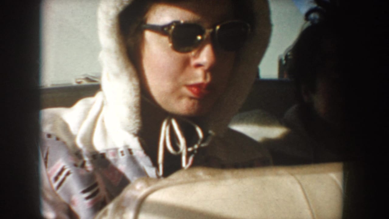 In a 1955 Idaho home-movie frame, a woman wearing dark sunglasses and a white hooded jacket sits behind a beige car seat.