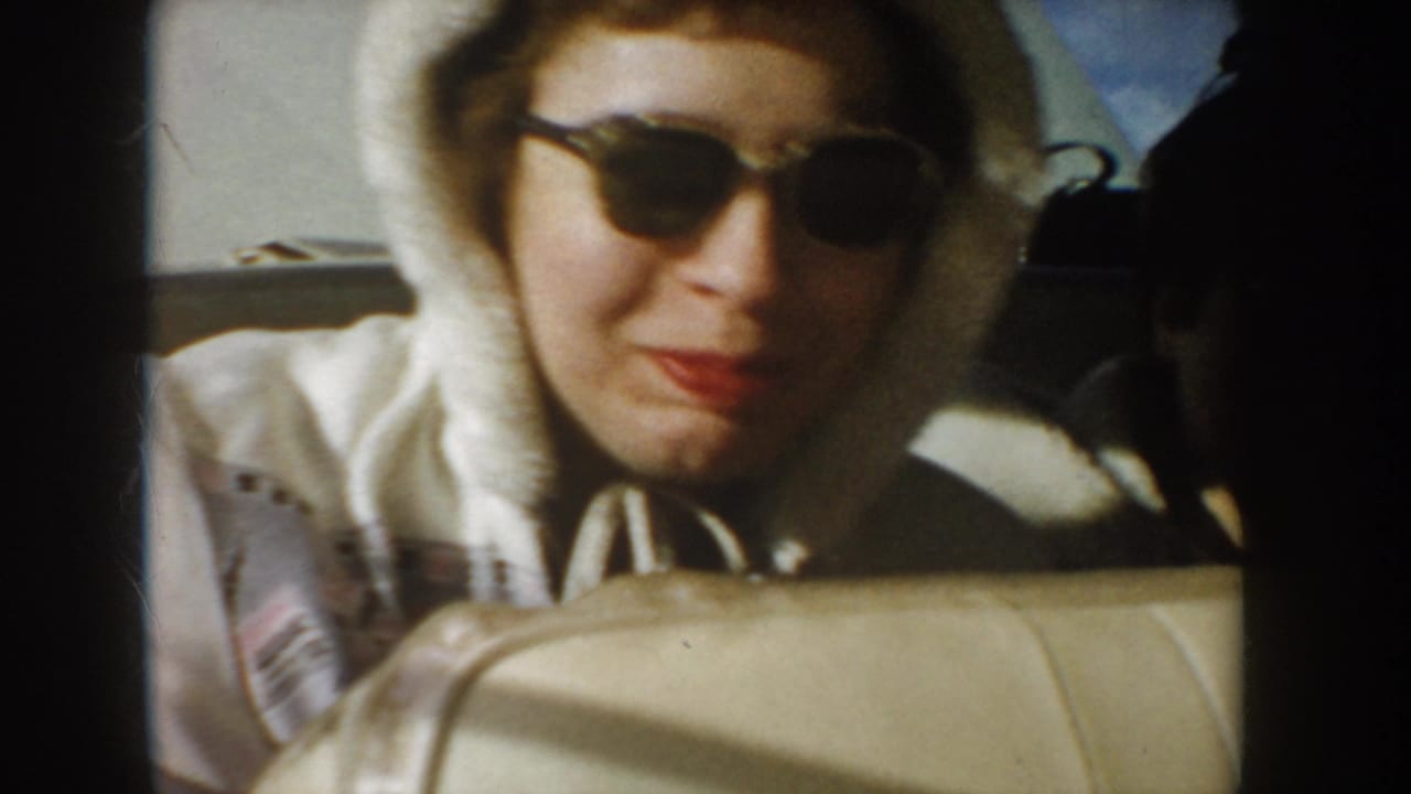 Woman in sunglasses smiling or speaking from behind a car seat.