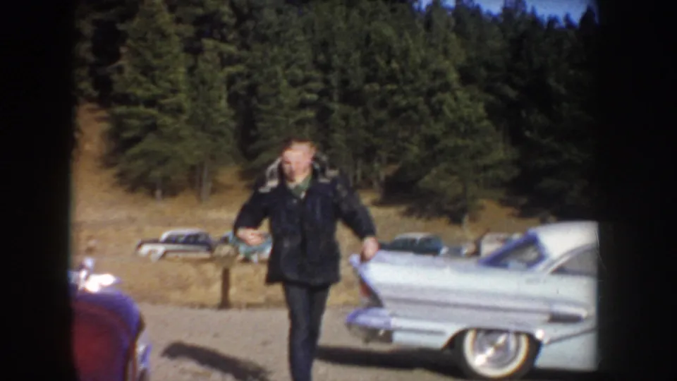 A man in a dark coat walks on gravel near classic automobiles, with trees and a hillside behind him in the supplied Idaho setting.