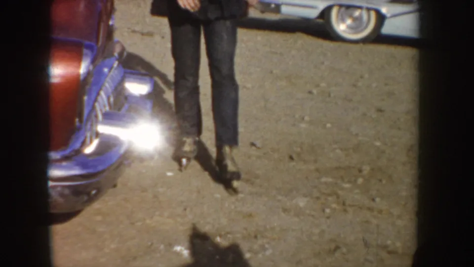 A cropped 1955 Idaho home-movie view shows a person's legs on gravel beside classic cars, with bright glare from a car bumper or light.