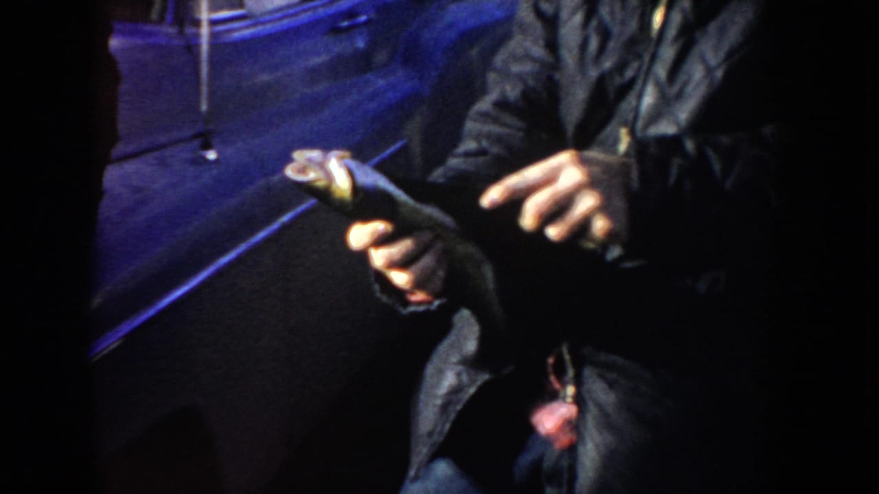 1955 Idaho 8mm frame showing a man’s hands holding a small fish near a dark car in blue night light.