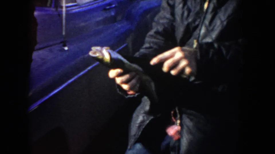 1955 Idaho 8mm frame showing a man’s hands holding a small fish near a dark car in blue night light.