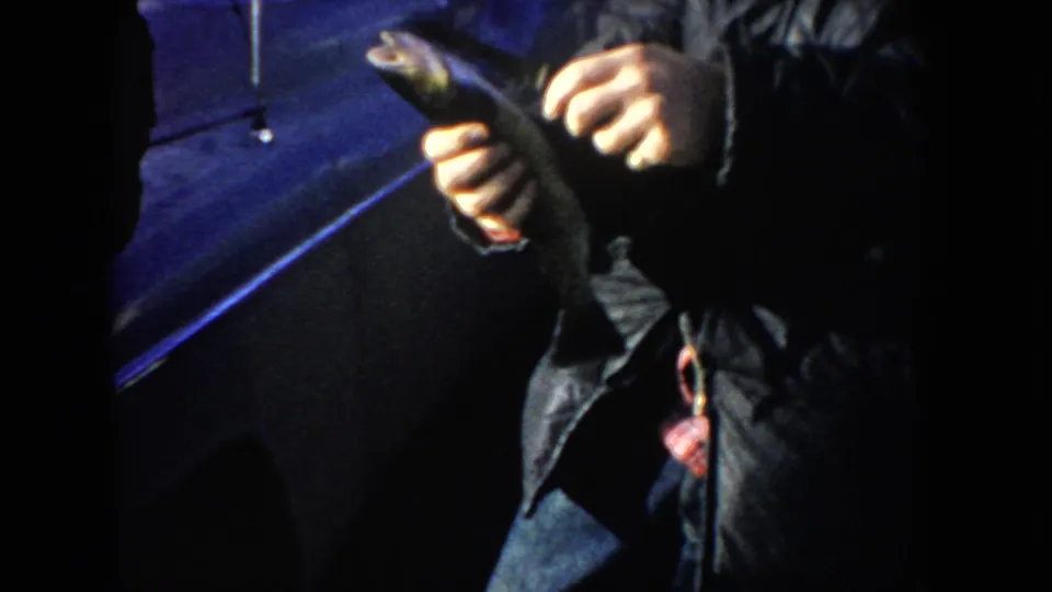 Close 8mm view of a man holding a small fish near a parked car, with the figure partly cropped by shadow.