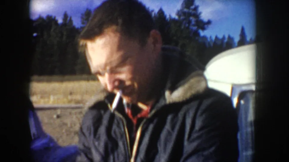 1955 Idaho home-movie frame showing a man’s face with a cigarette beside a car, with trees in the background.