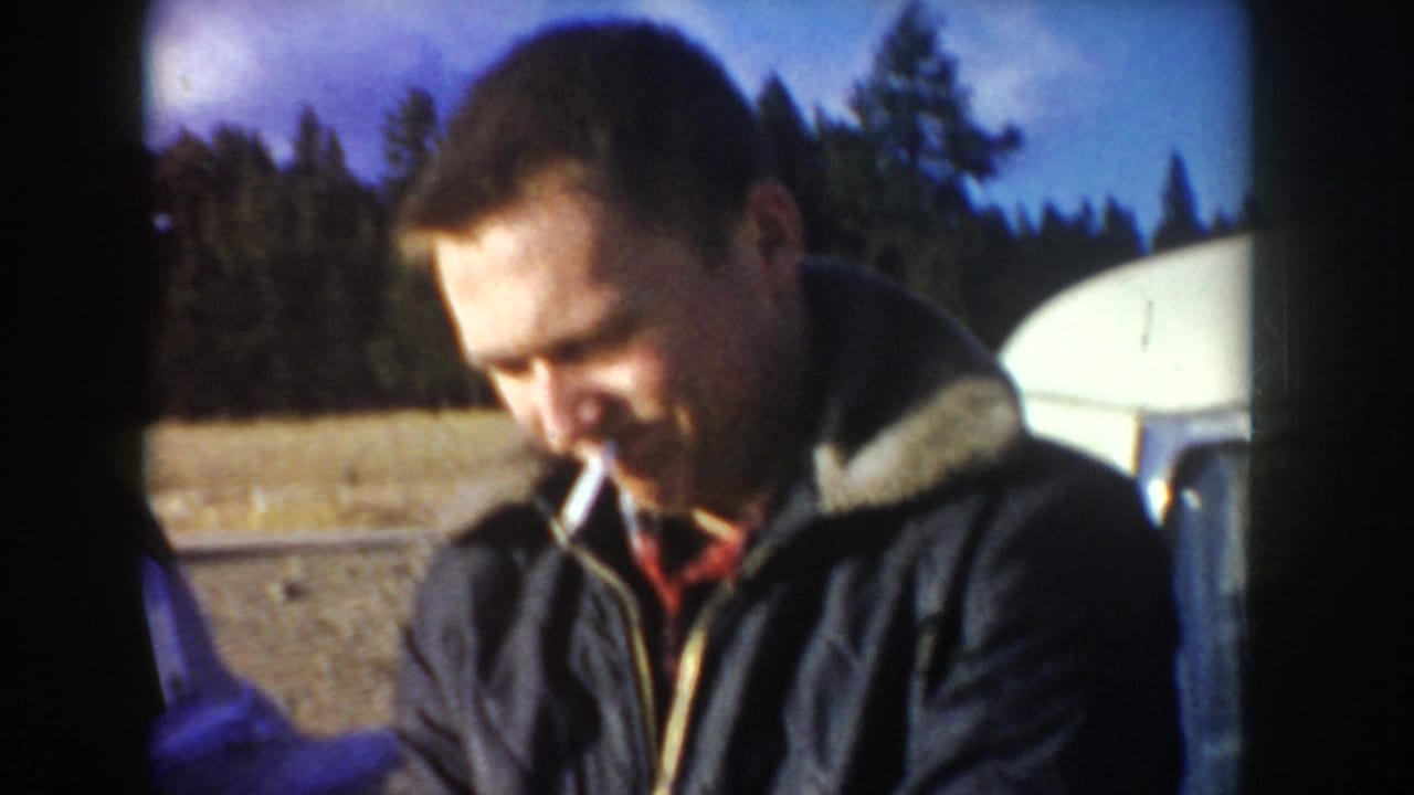 In 1955 Idaho home-movie footage, an adult man in a dark jacket smokes beside a white vehicle near a wooded outdoor area.