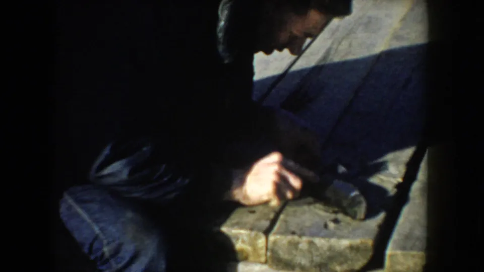 The man remains bent over the fish on the wooden dock, with strong shadow and soft 8mm color in the frame.