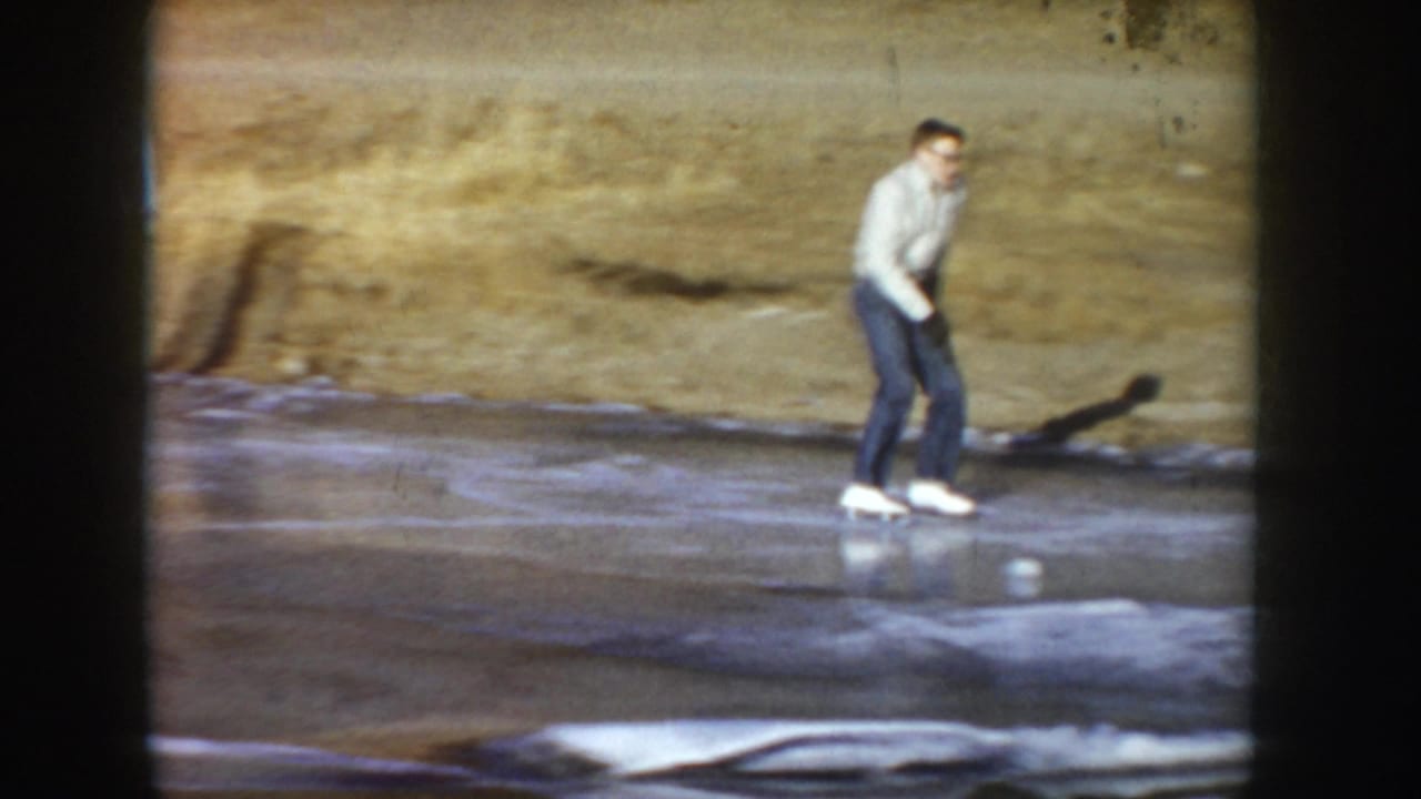 Young man skating on a natural frozen pond in what may be Idaho, circa 1955; white sweater, dark jeans, white ice skates; 8mm home movie frame.